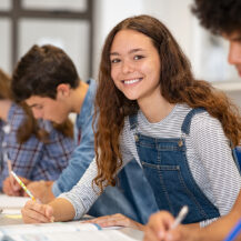 Satisfied young woman looking at camera. Team of multiethnic students preparing for university exam. Portrait of girl with freckles sitting in a row with her classmates during high school exam.